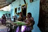 A woman speaks into a phone to search for information. She’s sitting alongside another woman — an internet ‘saathi’, or trainer — a table with a sewing machine on top, in front of a house with walls painted blue and yellow. Another woman watches on in the background.