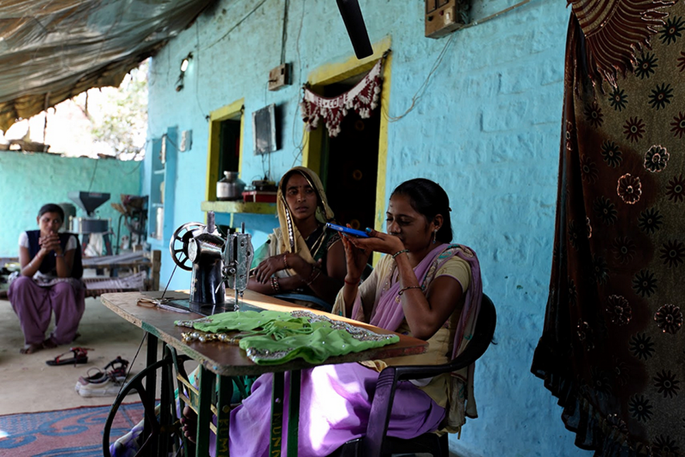 A woman speaks into a phone to search for information. She’s sitting alongside another woman — an internet ‘saathi’, or trainer — a table with a sewing machine on top, in front of a house with walls painted blue and yellow. Another woman watches on in the background.