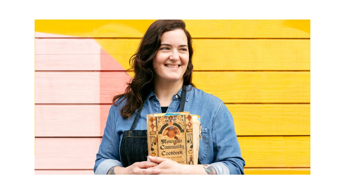 Jo Snyder holds a copy of her cookbook. She is looking beyond the camera, smiling. Her brown hair is down and over her shoulder. She is wearing a denim button-up shirt. She is standing against a yellow and pink wall.