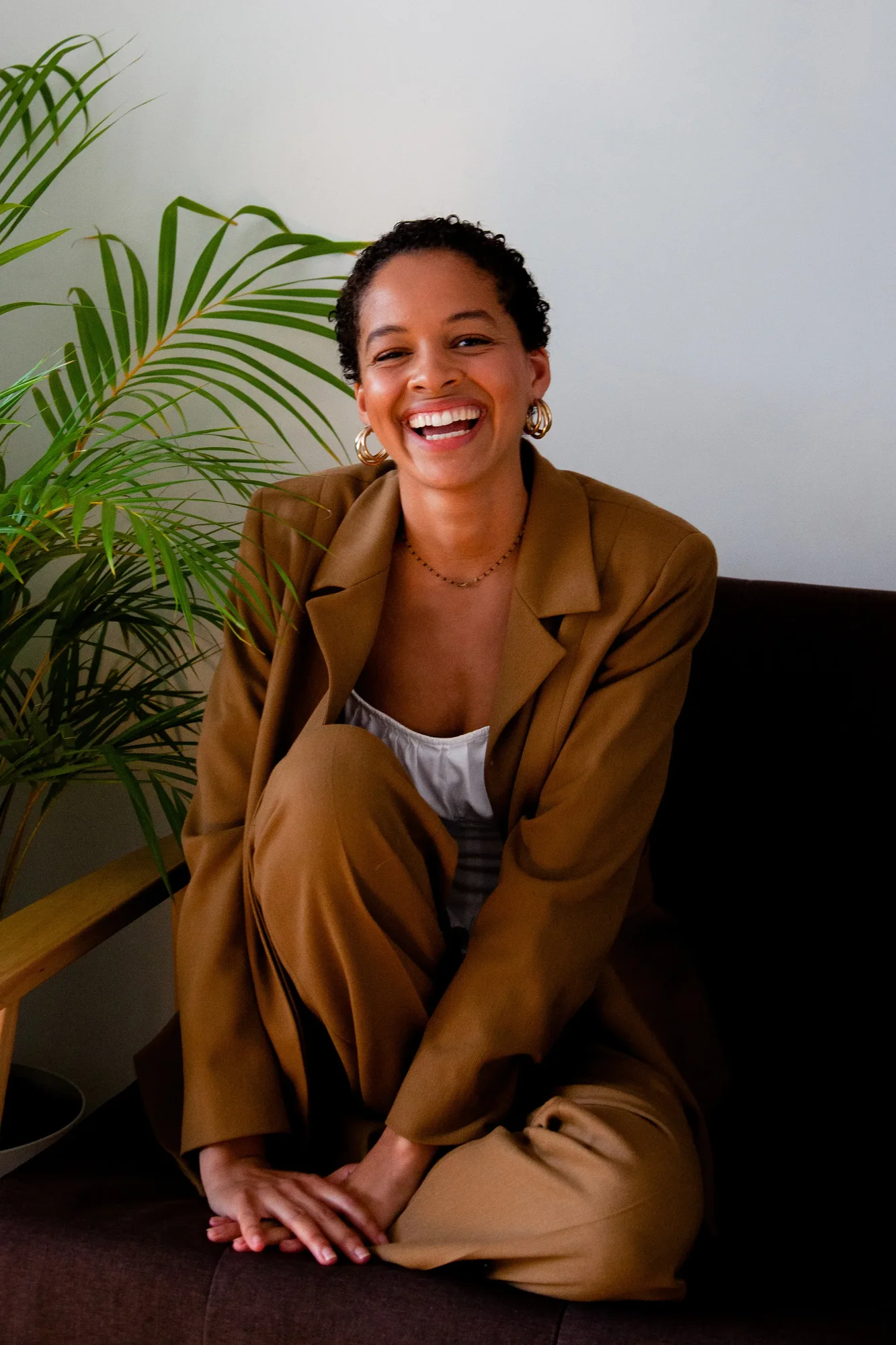 A woman wearing a brown suit and a white top sits crossed-legged on a dark brown couch, with a large green plant in background. She smiles at the camera and wrapped her hands around one propped-up knee.