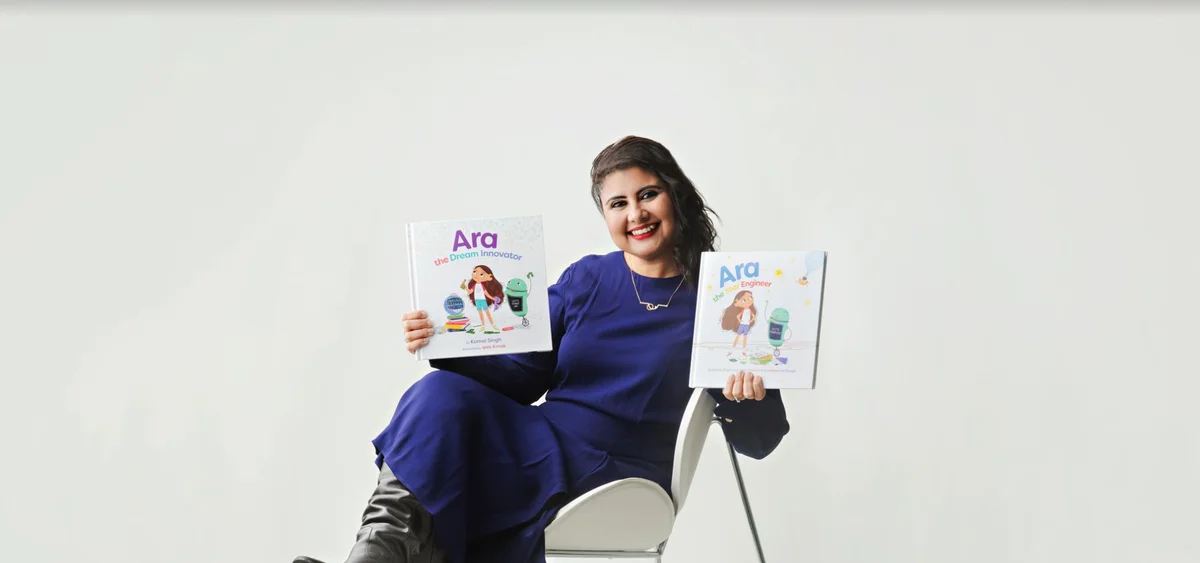 Photo of Komal Singh sitting on a chair against a white background. She is looking into the camera and smiling, holding a book in each hand. One is "Ara the Star Engineer" and the other is "Ara the Dream Innovator."