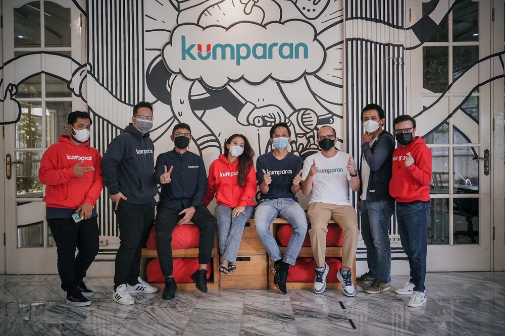 This is a photograph of the team at kumparan. There are eight people in the photo — seven men and one woman. A few of the team are wearing bright red hoodie sweatshirts with kumparan written on the front. Some are sitting down with others standing.