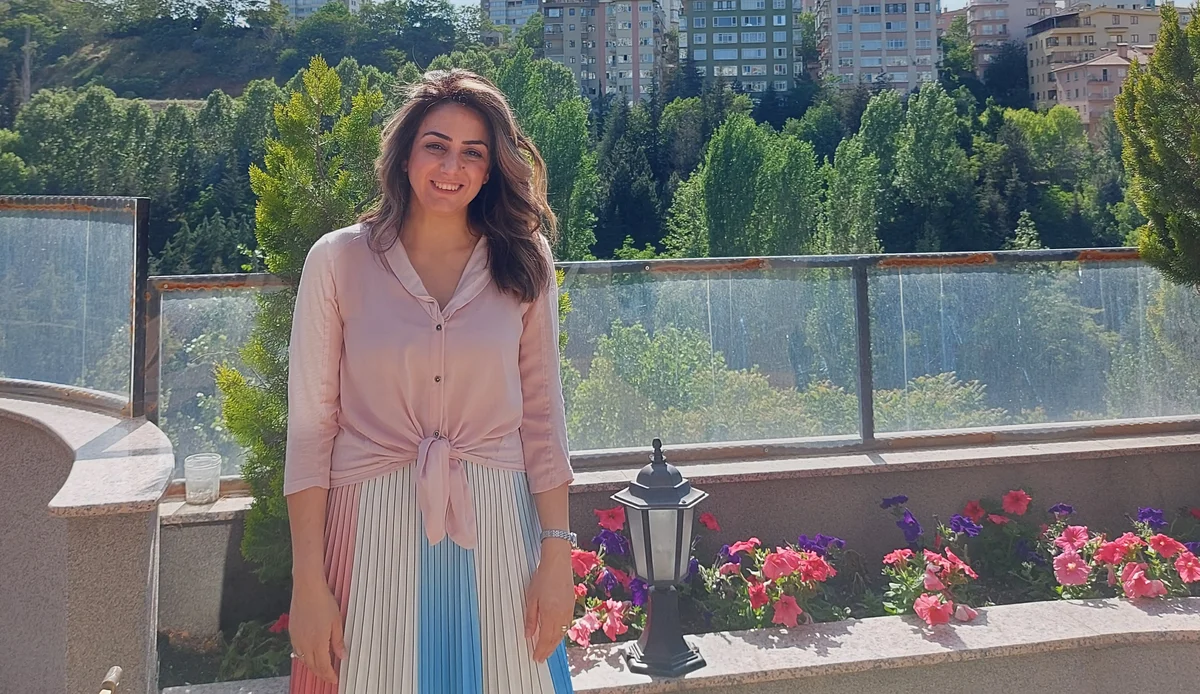 A woman with long hair, wearing a pink shirt, smiles at the camera with a city in the background