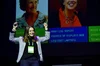 Lara presents at an event. She wears a gray jacket and a lime green event badge. Behind her are pictures of women technologists with their names and talk names listed.