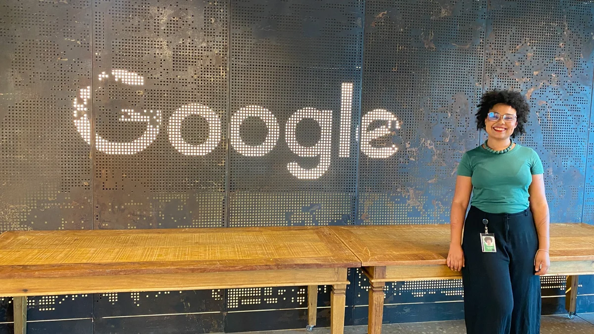 Larissa Botelho, mulher negra vestindo uma blusa verde e calça preta, posa em frente a mesas de madeira e um logo do Google ao fundo no ambiente do escritório em Belo Horizonte