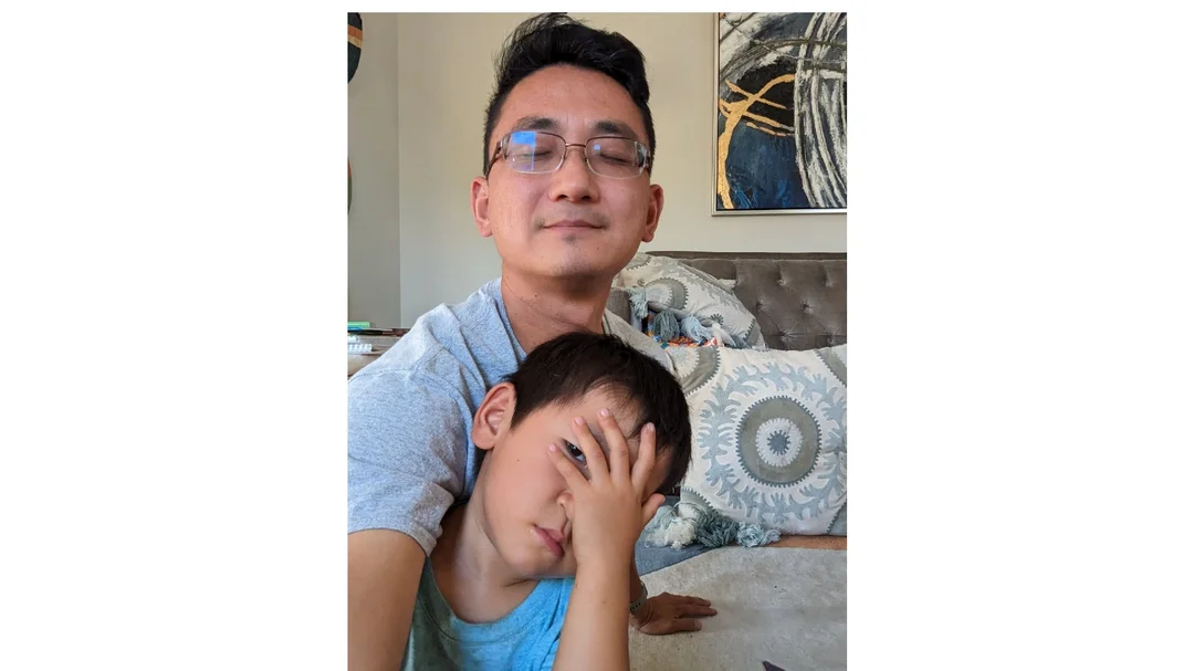 A selfie taken by a man who is using Guided Frame; his eyes are closed and he is smiling. A small boy who is covering his eyes and peeking out sits in his lap.