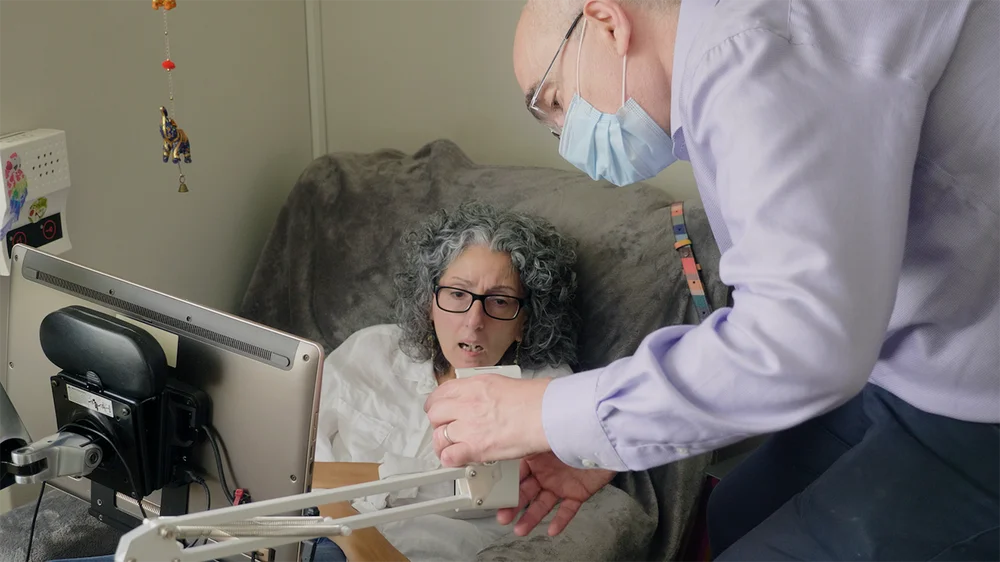 A man wearing a mask (Richard Cave) is seen chatting with a woman (Sarah Ezekiel) in front of her eye gaze tablet