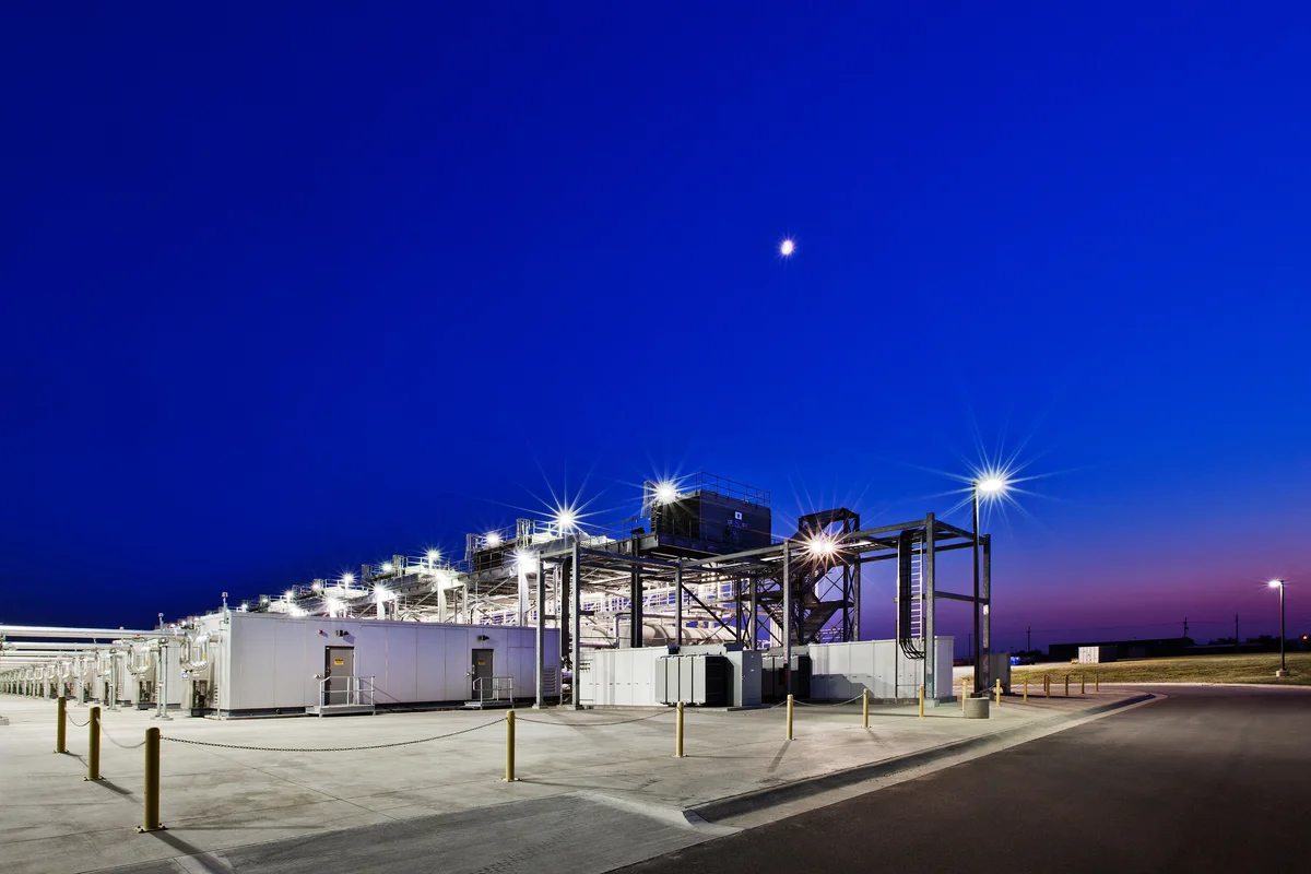 Bright lights shine on top of a line of white data center units, surrounded by metal scaffolding. As the moon shines above, the sky transitions from purple on the horizon to a deep blue.