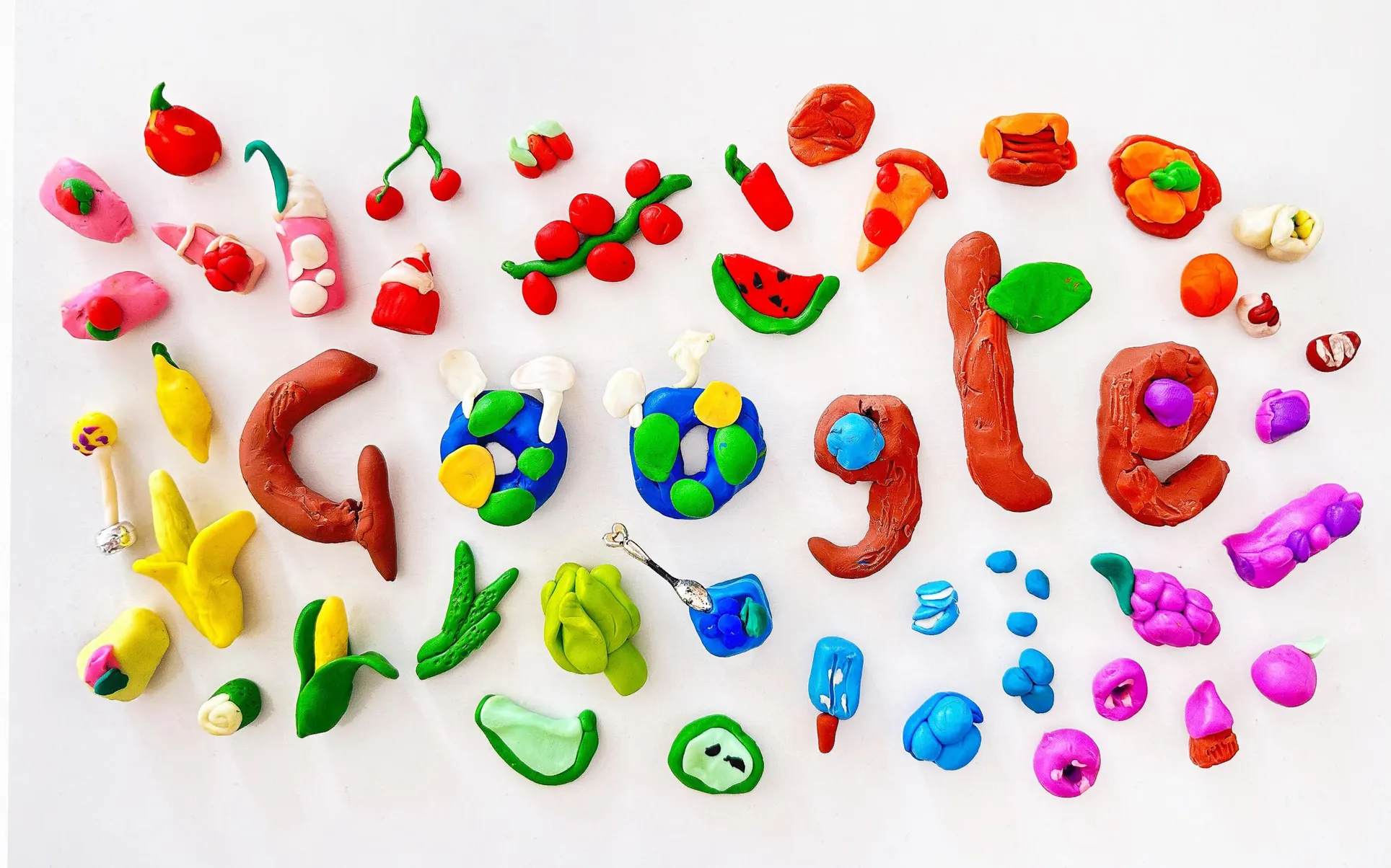 A clay sculpture depicting the Google logo surrounded by various food items. The letter G is formed from red clay. The Os are represented by blue two circles possibly meant to symbolize the Earth. The second G is constructed from red clay with a central blue clay disc. The L is fashioned from red clay with a green appendage, likely a leaf. Finally, the E is created using red clay with a purple clay disc.