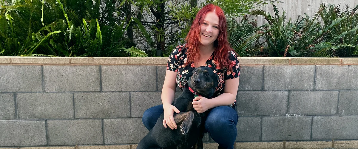 Nichole sits with a dog in front of a cement wall with plants on top.