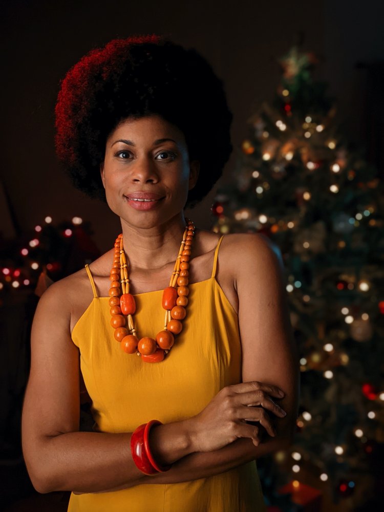 Image showing a woman in a yellow dress standing in a dark room in front of a decorated tree. The woman is well-lit and in focus while the tree in the background is blurred.