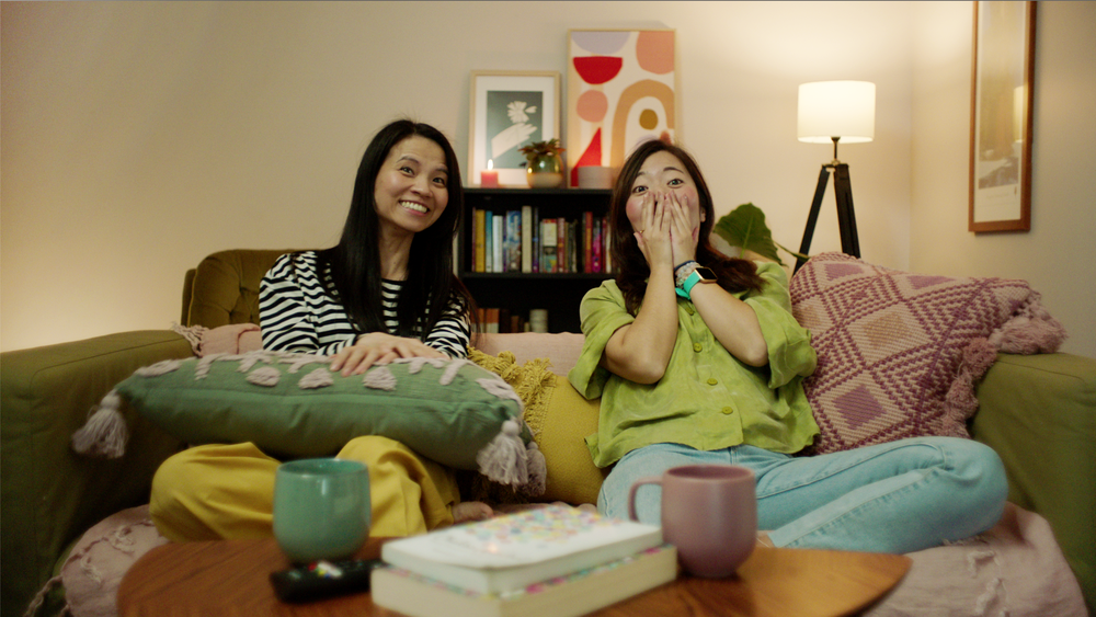 Two friends are lounging on a couch while watching a TV screen. One is smiling and the other has their hands covered over their mouth in surprise.