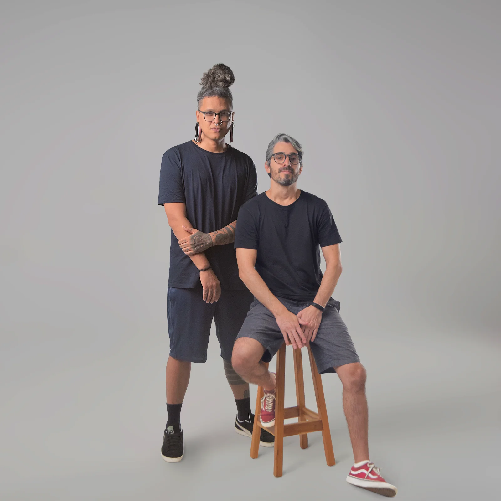 Two people wearing all gray clothing look into the camera. One of them stands with one arm crossed, the other sits on a wooden stool.