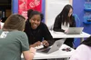 Image shows two women sitting at a white desk talking over an open laptop. One woman has her back to the camera and is looking at the laptop screen, the other women is facing the camera, looking at the other woman, and smiling.