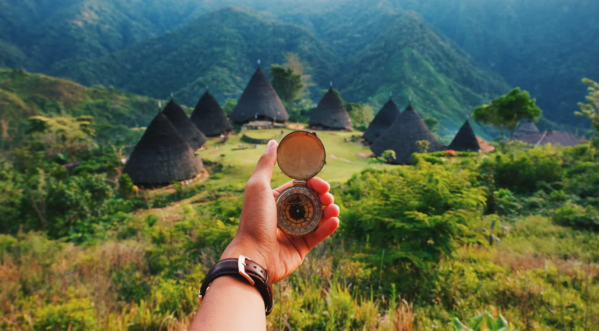 A left hand extends into the photo holding a compass — there are lush green hills and a village made up of large straw structures in the background.