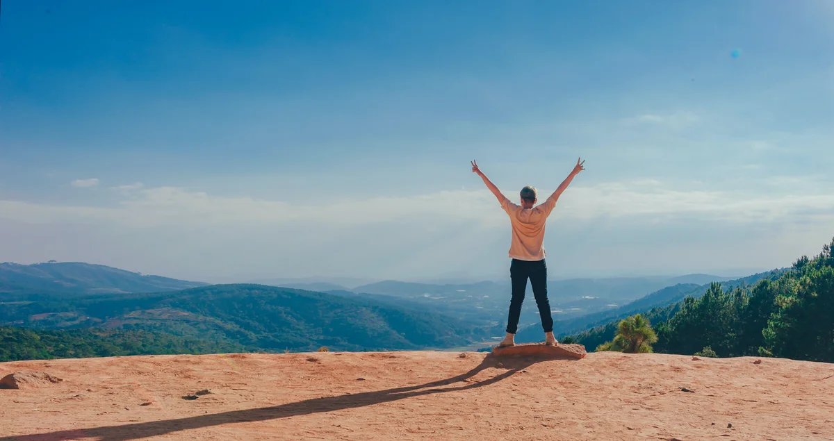 A person standing on a mountaintop, back to the camera, with arms outstretched, looking at a view of nature