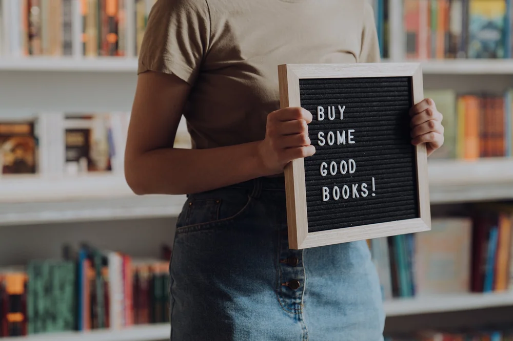 A woman standing in front of a bookshelf holding a sign that reads "BUY SOME GOOD BOOKS!"