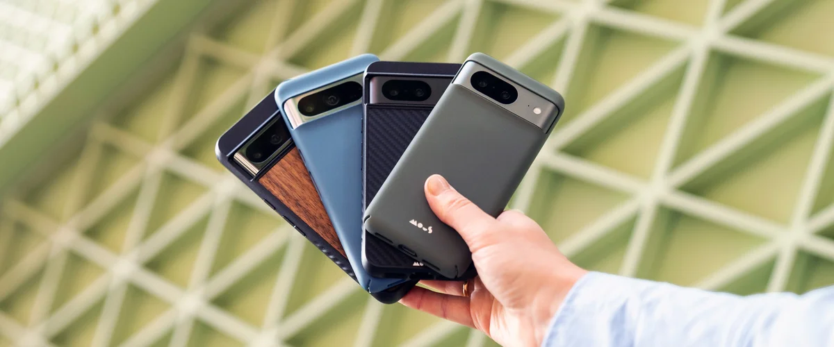 A hand holds four phones fanned out: One has a wood detailing case. The second has a blue case. The third has a dark blue case with angled lines etched in it. The last has a gray case with a small logo at the bottom that reads “Mous.”