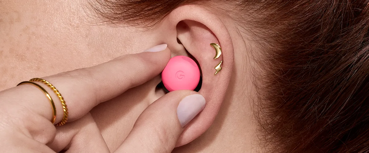 Photo of a close-up of the side of a person’s head, revealing their ear featuring two small golden earrings and their hairline behind their ear. Their hand is reaching to place a pink Pixel Bud into their ear; the Google “G” icon is visible on the front.