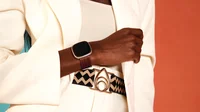 A woman in a white pantsuit and geometric belt modelling the new Brother Vellies woven leather accessory band in oak for Fitbit Sense 2 and Versa 4.