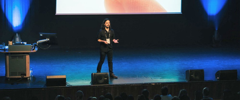 Ricardo stands on a dark-lit stage in a black shirt and black pants, and presents to an audience. He is standing next to a podium and in front of a presentation screen.