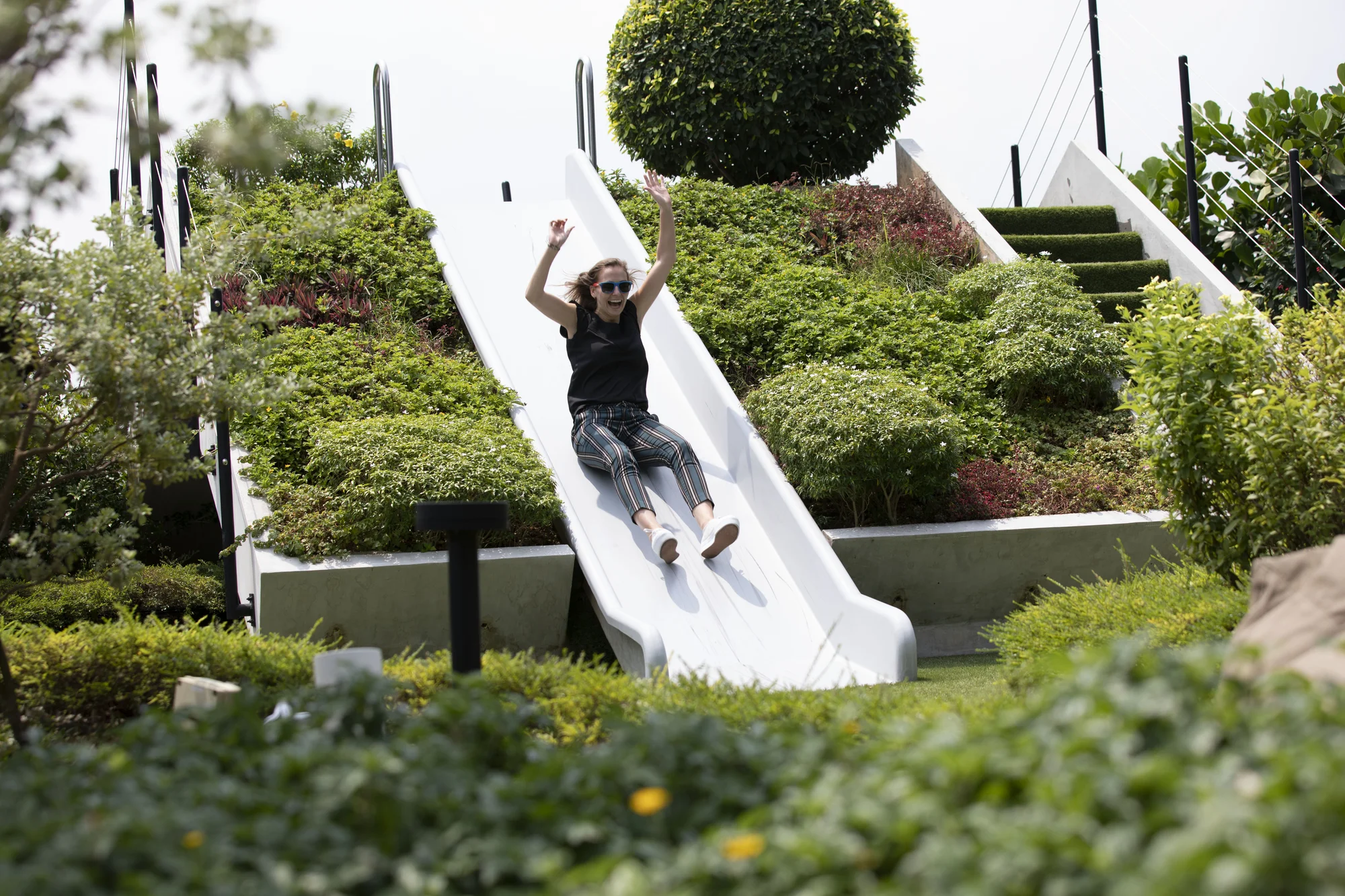 A woman raises her arms as she slides down a slide. Around her are plants, planters and a staircase through the garden.