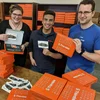 One woman and two men stand looking at the camera, holding orange boxes that say "Thimble" in white letters. More orange boxes are on the table in front of them as well as on the shelves behind them.