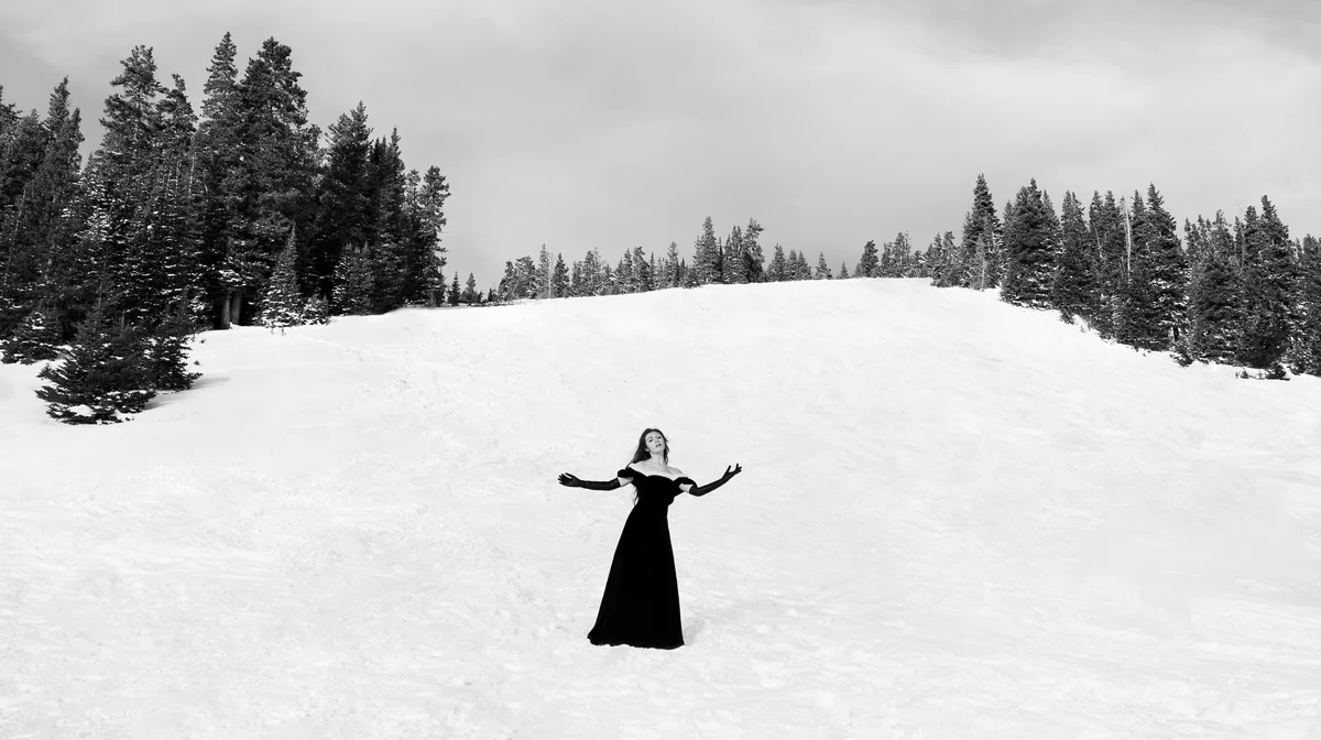 Black and white photography of a woman in a black dress standing in the snow. There are trees in the background.