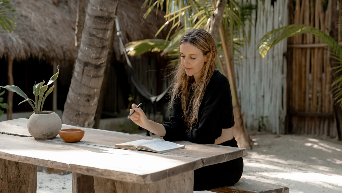 ArticlWearing a black dress and seated at a wooden table in a tropical setting, wellness blogger Andi Eaton writes in a notebook. e's hero media