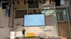 Hannah stands on stage, speaking into a microphone. In the background is a screen showing presentation slides, mounted on a white wall with the words “Further Faster Together” on the right side, and list of large cities on the left. In front of the wall is a yellow couch and grey couch, and a podium with the Google for Startups logo on it.