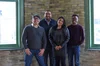 Three men and one woman stand looking at the camera in front of a brick wall .