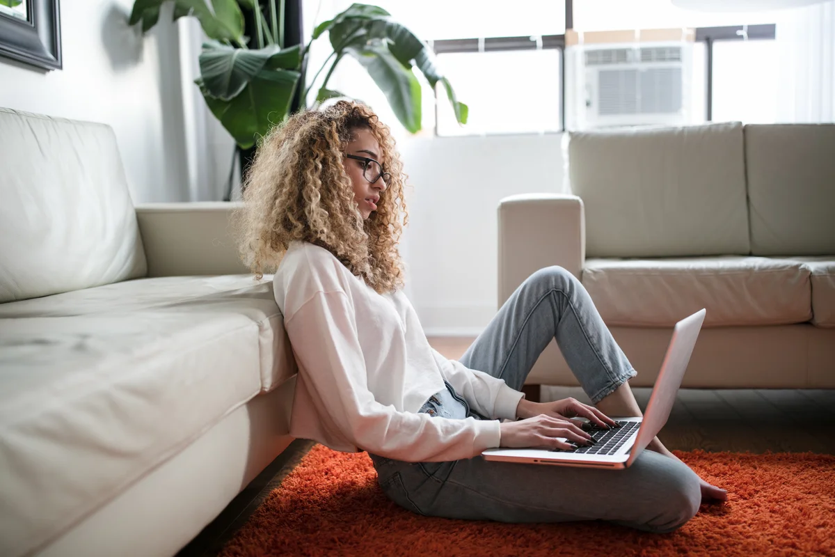 Mulher sentada no tapete da sala de estar, encostada no sofá, trabalha no notebook.