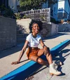 Tiffany Williams sits on a curb painted blue in a white shirt, brown shorts and white boots.