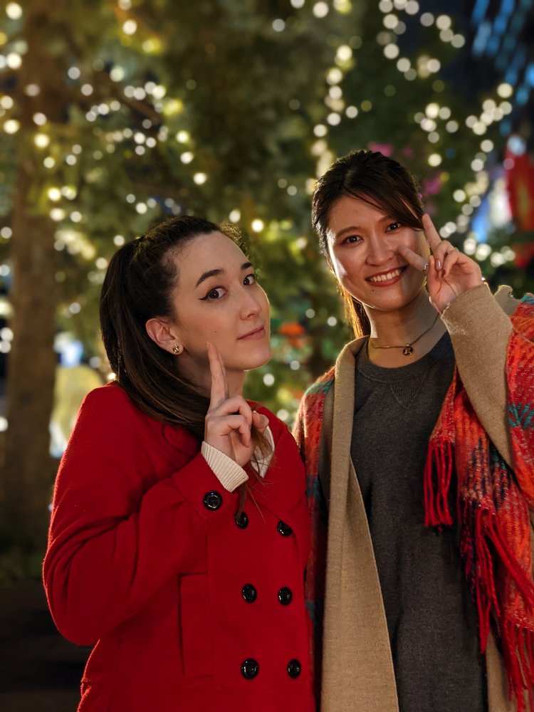 Image showing two women smiling at the camera. They are in focus while the decorated tree in the background is blurry.