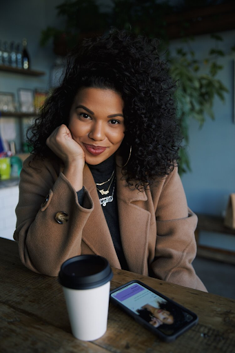 Tyla-Lauren smiles as she sits at a wooden table with a hot drink and phone in front of her. She wears her hair long, in curls, with a beige coat and black shirt.