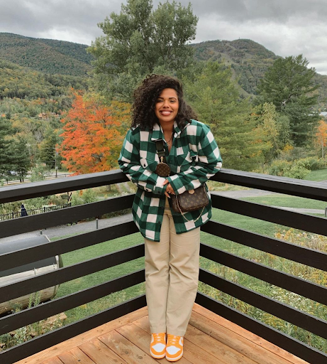 Tyla-Lauren stands on a deck outside with a checkered flannel shirt. A mountain and trees with changing color leaves are in the background.