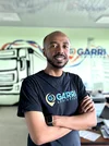 A man wearing a black shirt with blue and yellow "Garri Logistics" logo stands in an office with his arms folded looking at the camera. Behind him is a mural of a truck.