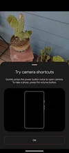 A screenshot of a Pixel display showing the camera open to photograph cacti in pots on a porch. A pop-up black boxed prompt takes up half the screen and reads “try camera shortcuts. Quickly press the power button twice to open camera. To take a photo, press the volume button.” Below this is an outline of the shape of a Pixel phone including the side buttons. Underneath this is a pill-shaped box that reads “OK.”