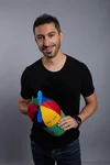 Ernesto, in a black T-shirt and blue jeans, holding his Noogler hat and smiling to the camera.