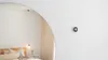 A photo of a white wall with a portion of a circular mirror in the left third of the image. The mirror is reflecting a bedroom, showing part of a bed, a hanging light, and a curtain. On the wall next to the mirror is a silver Nest Thermostat.