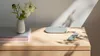 A photo of a pill-shaped, gray-green Google TV Streamer on a light wood surface. It’s next to a pill-shaped remote of the same color. A small stack of books with a white vase of flowers sits on top of the books. Shadows stream across the objects.