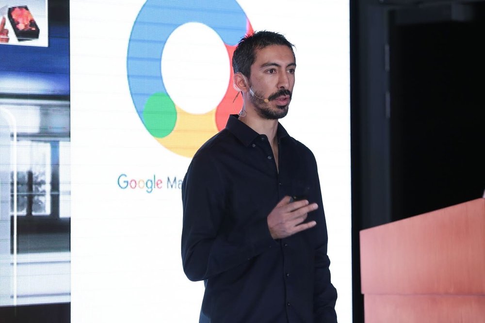 Ernesto, in a dark blue button down with the top two buttons undone, speaking at a Google marketing event.