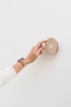 A photo of a hand wearing a gold Pixel Watch reaching out with one finger to touch a gold, circular Nest Thermostat against a white wall. The thermostat reads 78 degrees.