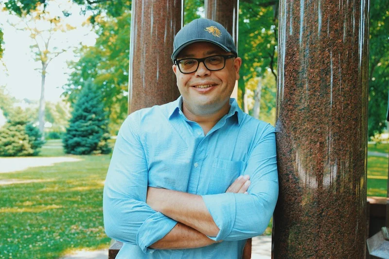 A man wearing a blue shirt and baseball cap leans against a stone column in a park.