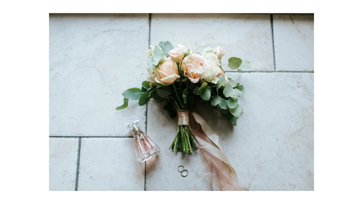 Photograph of a wedding bouquet next to a small bottle of perfume and two rings.