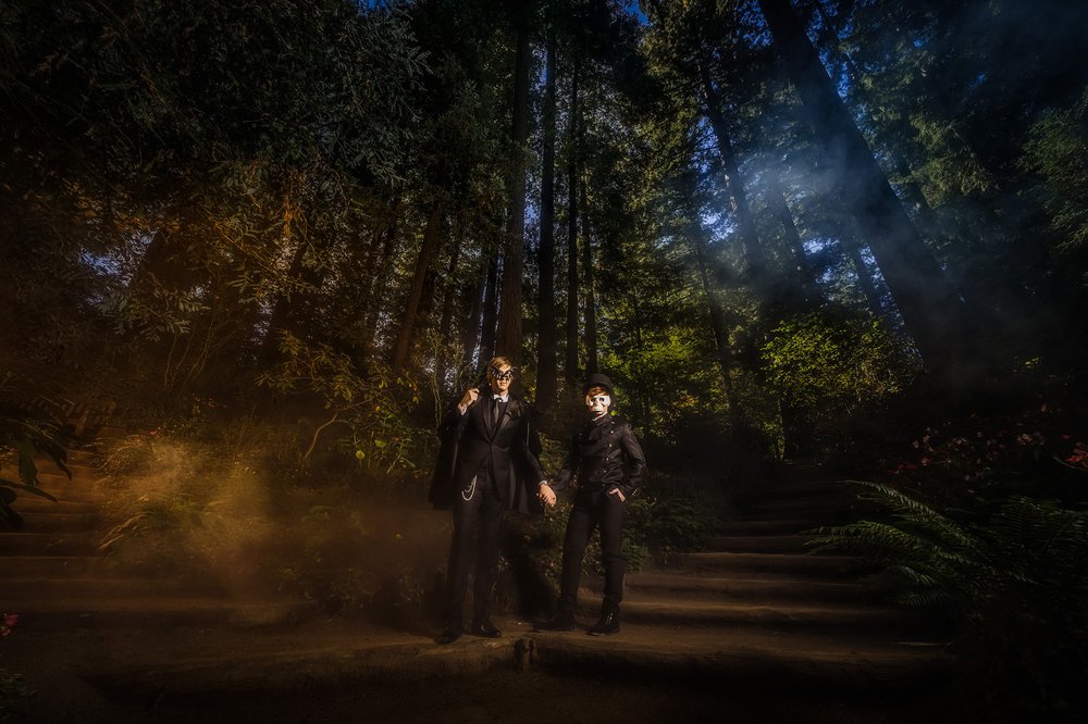A masked couple in head-to-toe black suits stand side by side in a dark forest. Photo: Wild About You Photography