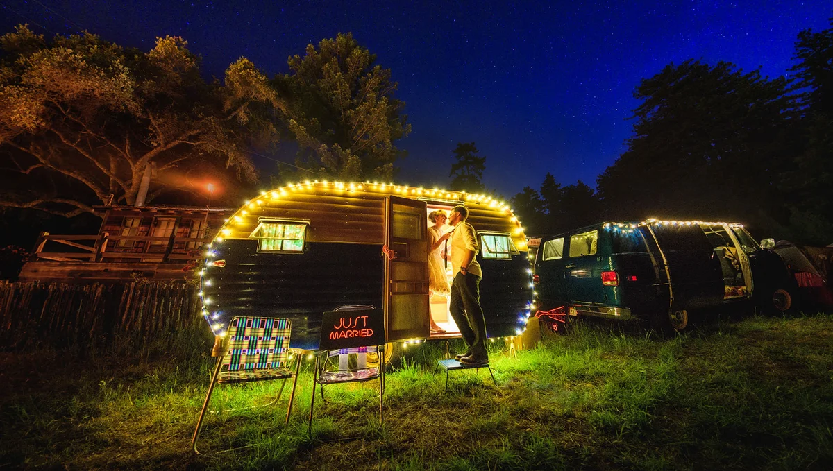 A bride and groom gaze at each other in the open doorway of a camper lit by string lights, set in a grassy field. A sign set on a folding chair outside the camper says “just married.”
