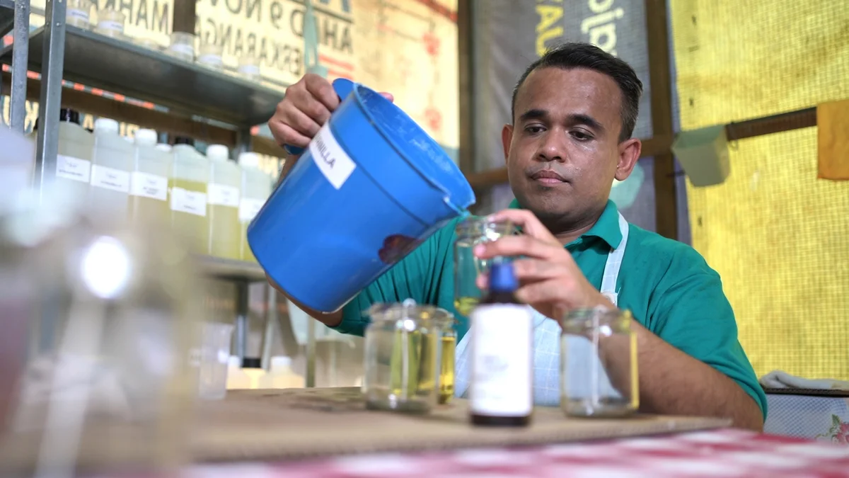 Business owner Mohd Zaid — wearing a green shirt and white apron — pours oil from a blue bucket into a glass jar. There are a large number of jars on the table in front of him, and many jugs of oil on the steel shelving behind him.