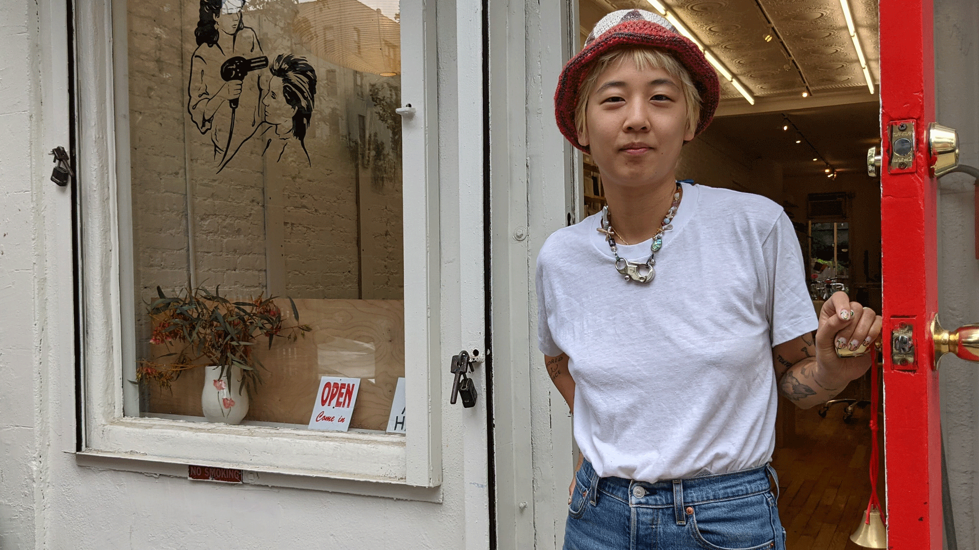 A person standing in front of a white business store-front. They’re wearing a white shirt, blue jeans, a red hat, and a necklace welcoming a customer into the shop.