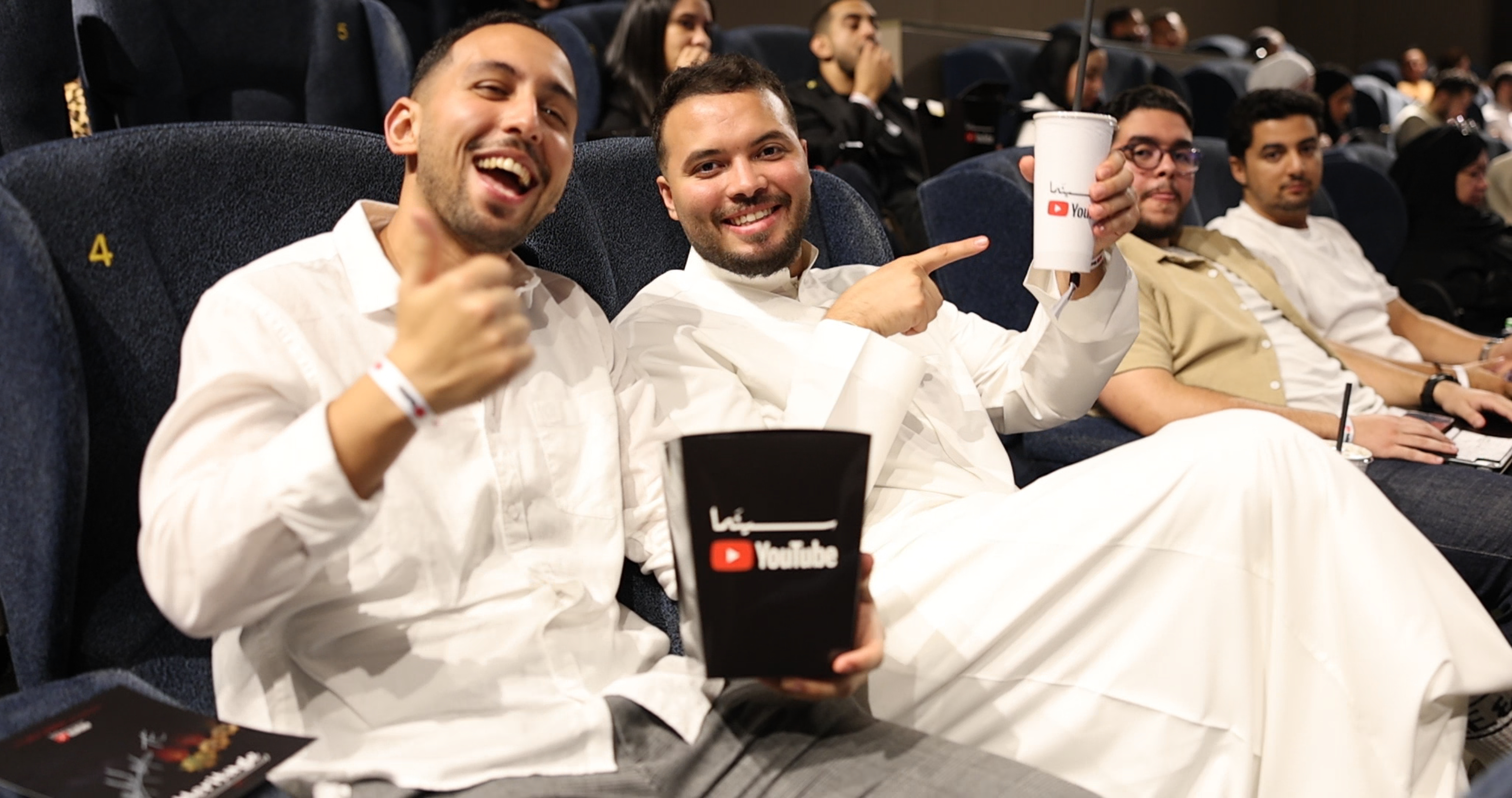 Two young men sit in comfortable cinema seats, smiling at the camera. The man on the left wears a white shirt and gray trousers, giving a thumbs-up. The man on the right wears traditional white attire and points his index finger at the camera while holding a white cup with a YouTube logo. Between them, a black popcorn box also bears the YouTube logo. In the background, other people sit in cinema seats, some looking at the screen and some at the camera.
